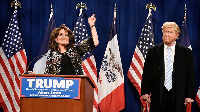 Tina Fey stands at a lectern with a "Trump" sign on the front as she plays Sarah Palin, US flags i a row behind her. Darrell Hammond as Donald Trump is to her left, giving her the "side eye" look.