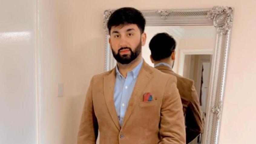 A Pakistani man with short brown hair, beard and moustache. He is posing for a picture in front of a mirror and is smartly dressed in a blue shirt and tan suit jacket.