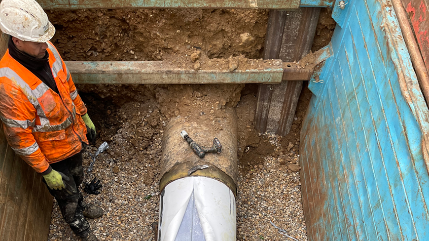 A contractor wearing an orange high-vis jacket and a white helmet stands into a pit next to an exposed pipeline, with a white tube which fills the pipe being fed into it.