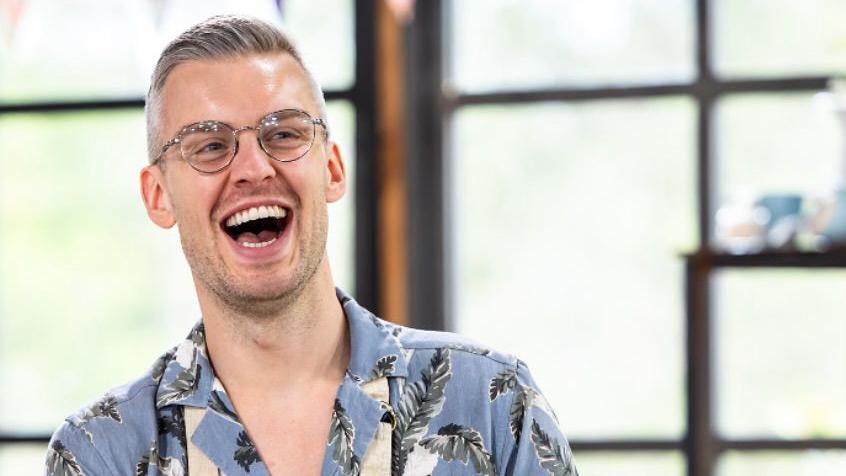 Elliot Styche laughs as he stands in front of a window during filming for The Great Australian Bake Off. He has short, slicked-back grey hair and wears glasses and a blue shirt with a grey and white fern-leaf design.