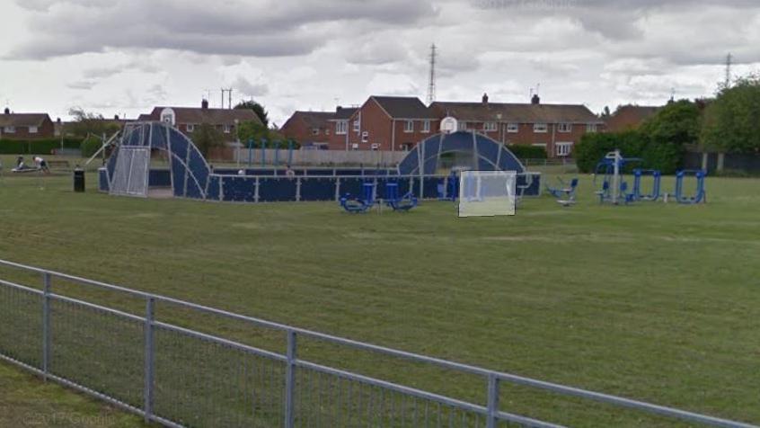 View of blue and grey-coloured play equipment at the park, with grass and metal railing in the foreground