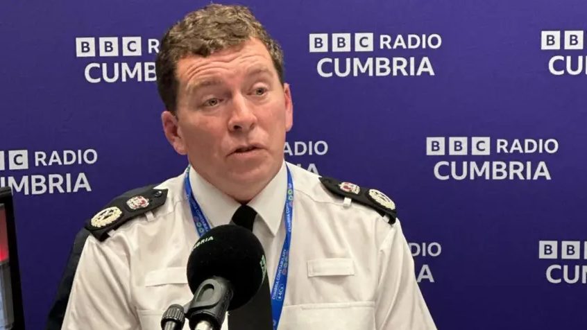 A wide shot of Rob Carden, in his police uniform. He has curly brown hair and is sitting in front of a black microphone to be interviewed for BBC Radio Cumbria.