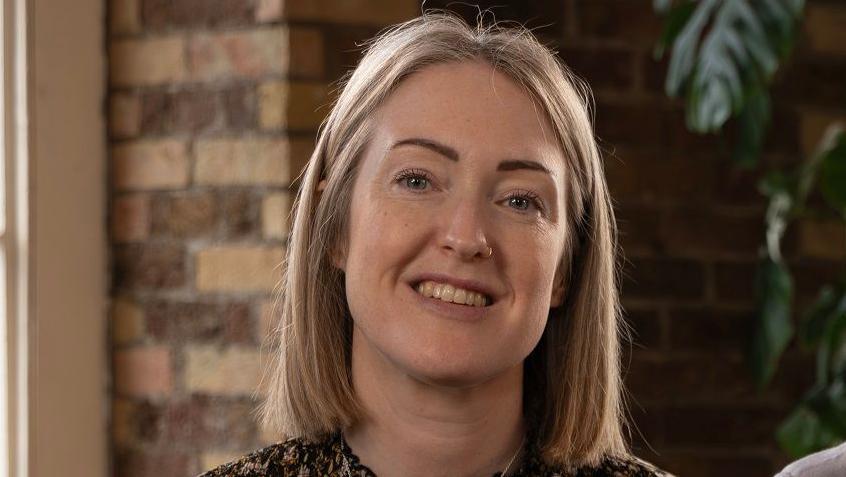 Esther Ghey, who has straight, shoulder length blonde hair, smiles for the camera. She is photographed in front of a brick wall and a large green plant.