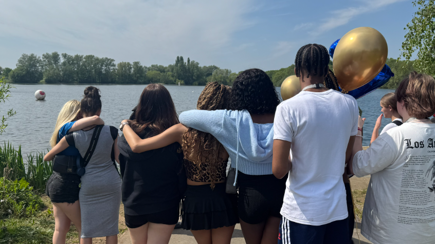 A group of young people looking out towards a lake. One holds blue and gold balloons