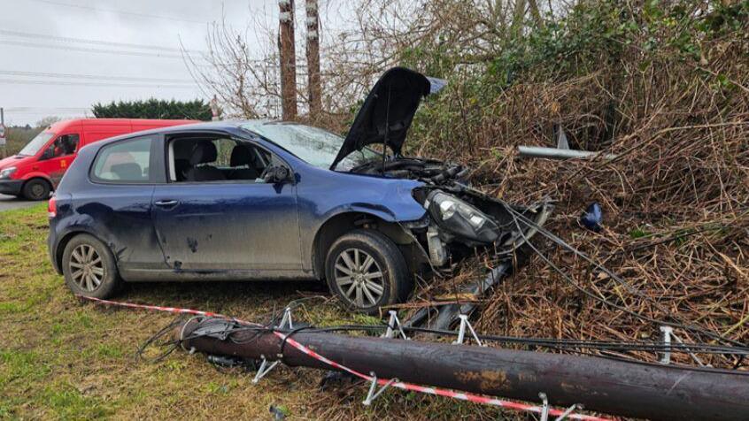 The scene of a crash in which a car has collided with a telegraph pole and come to a stop in a hedgerow next to the road. The car is a small, dark blue hatchback, with the front completely crumpled and the bonnet lifted. A large dark brown telegraph pole is visible, knocked down by the crash. There is lots of debris near the car. 