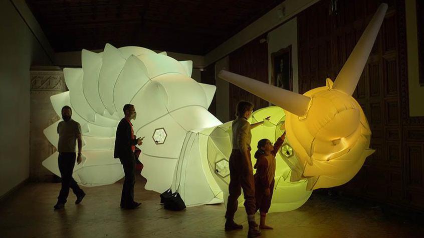 Huge illuminated inflatable snail looms above four visitors in a darkened museum hall as a child reaches out to touch the yellow head. 
