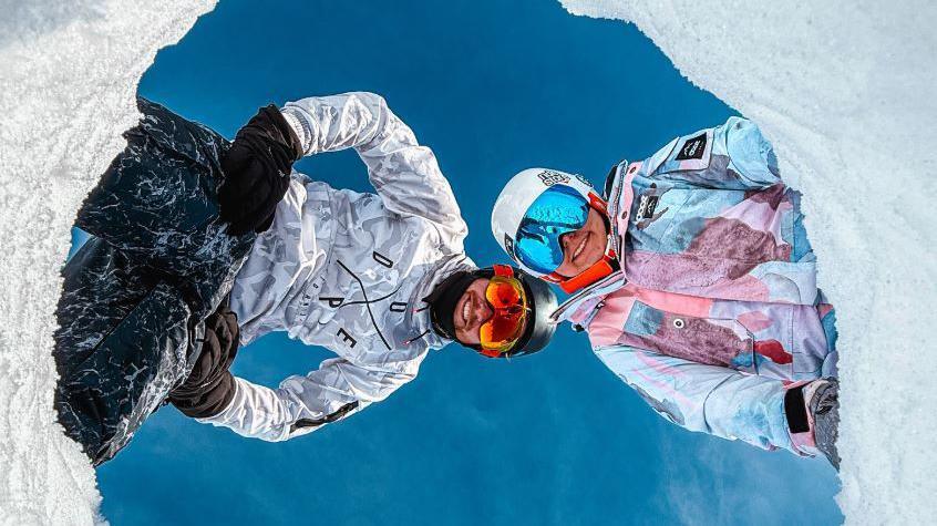 Hannah Bird and Charlie Camper wearing full snow suits, helmets and ski goggles. They have dug a hole in the snow and placed their phone in there, pointing upwards to capture them looking down against the bright blue sky.