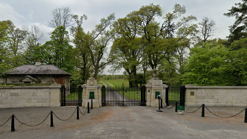 The gated entrance to hotel at Ballyfin Demense.  There is a stone wall with black iron gates, a gatehouse and a long laneway through a large lawned estate.