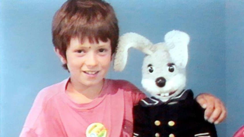 A young Andrew Guttridge wearing a pink t-shirt with his left arm round a Gus Honeybun puppet while visiting Flambards.