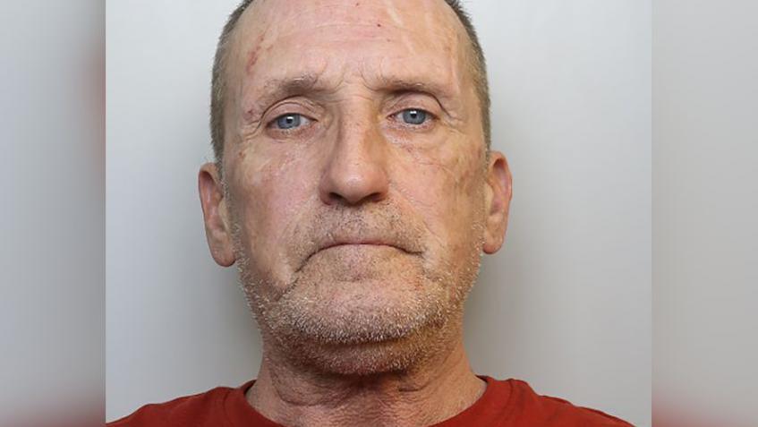 Stephen looks at the camera with a blank expression. He has short grey hair, a stubbly beard, and wears a red T-shirt.