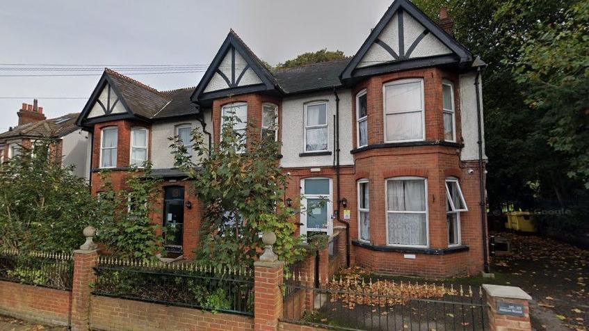 An exterior view of the suburban property, a large red brick building with the upper storey pebble dashed, white PVC window sills and shrubs and bushes in full bloom in the garden, behind a set of Victorian iron railings.