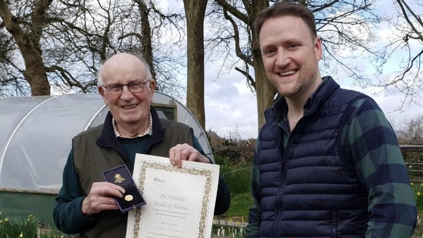 Two men are standing in an allotment. The man on the left is holding a certificate in one hand and a medal in the other hand. To his right is a man wearing a blue body warmer and chequered shirt. They are both smiling at the camera.
