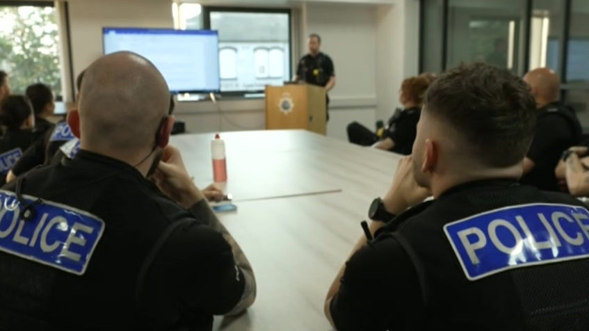 Several officers sit in a room with their backs to the camera, listening to another officer standing by a screen inside an office. The officers are in uniform and sitting around a grey desk.