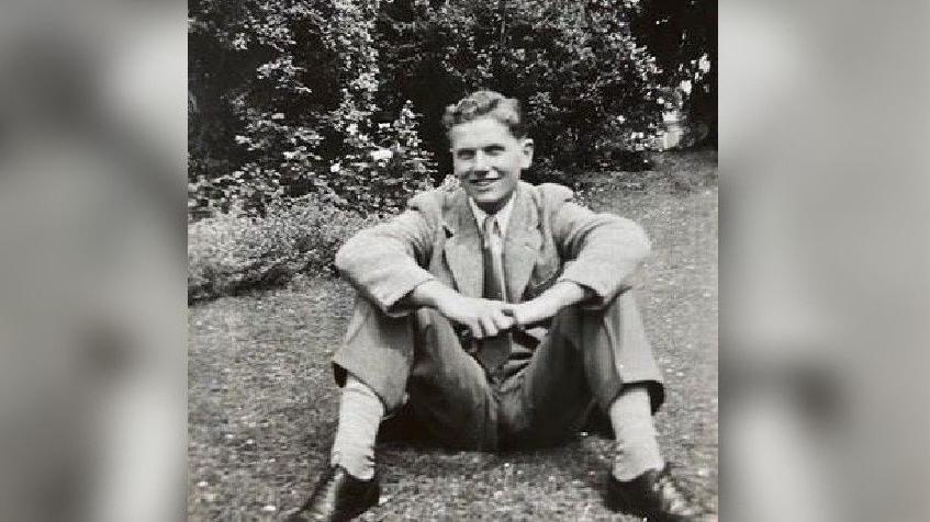 A black and white picture of Gordon wearing a light shirt, tie and smart trousers with a light coloured over blazer. He is wearing black shoes and light socks and is smiling at the camera, with trees behind him. It was taken in his garden in 1951.