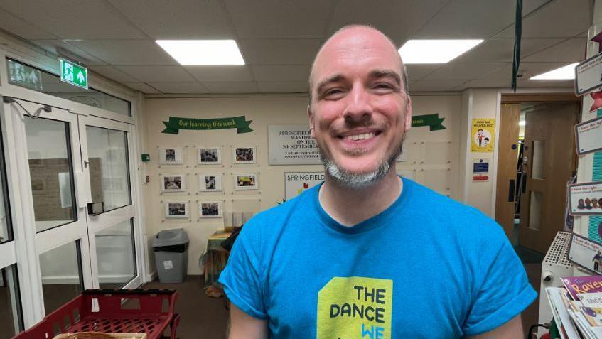 Tim Casson in a classroom wearing a blue t-shirt. He is smiling.