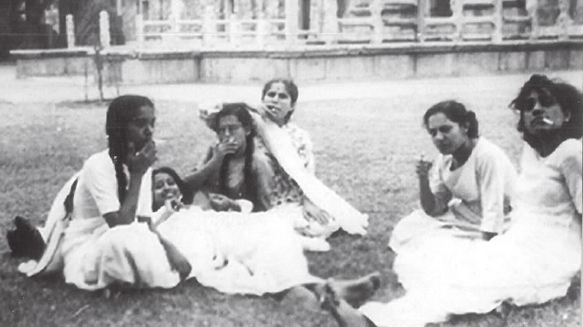 A black-and-white photo of a group of women sitting on the grass and smiling at the camera. Some of them appear to have cigarettes in their hands.