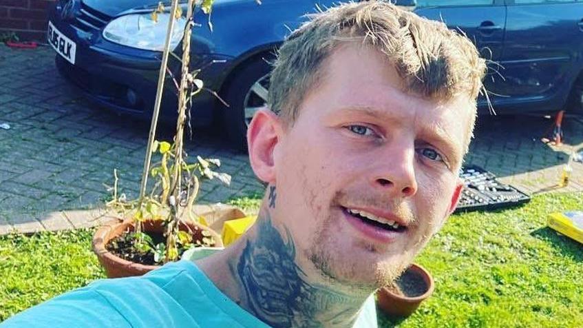 A man with short blond hair, a tattoo on his neck and a pale blue top in a grassy area with a dark blue car in the background