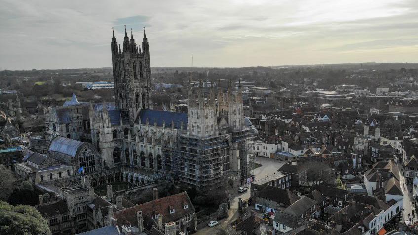 Canterbury Cathedral, which is a large, stone cathedral in gothic style. It is partly covered with scaffolding and towers over the surrounding city of Canterbury.
