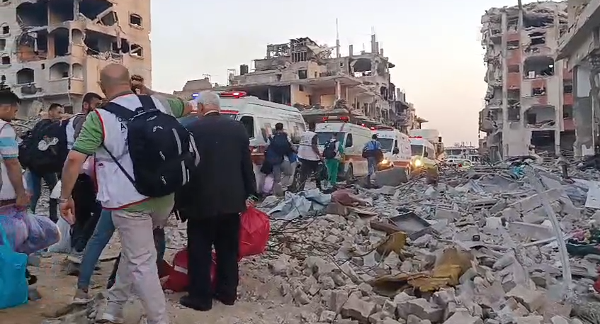 People wearing vests with the name of al-Awda hospital on the back carry bags and items to a row of ambulances waiting between the rubble of buildings
