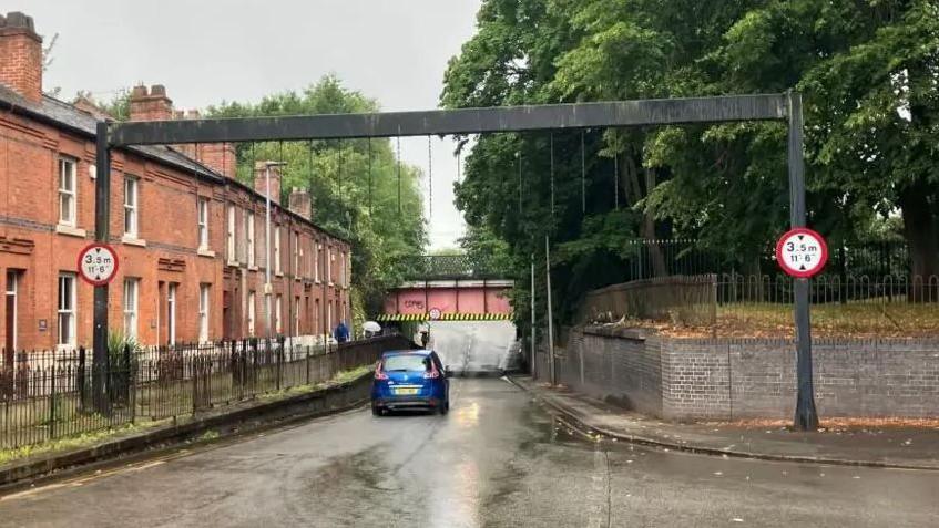 A blue car drives under a canal bridge on a residential road. People walk along the left side of the road with umbrellas.
