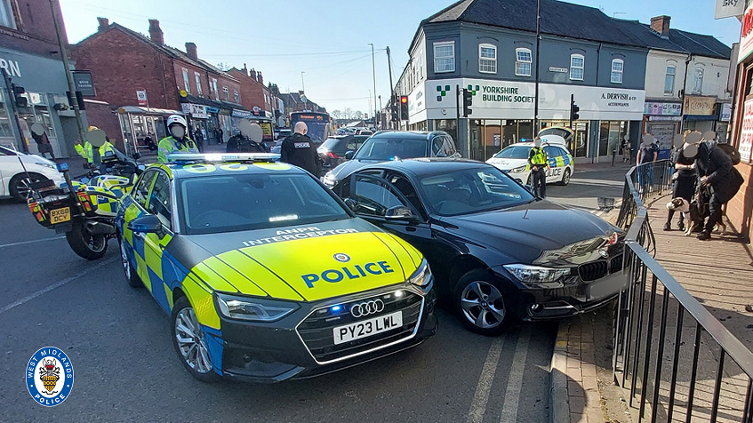 A police car which says 'interceptor' on it is parked diagonally across a road next to a black car which appears to have driven into black railings on a pavement. A police motorbike and police officers are behind the cars, which are on a busy road with shops.