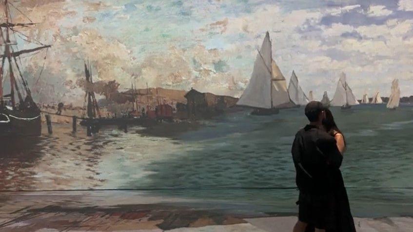 A young couple dressed in black stand to view a projection of one of Claude Monet's paintings, in which sailing ships are depicted in a harbour.