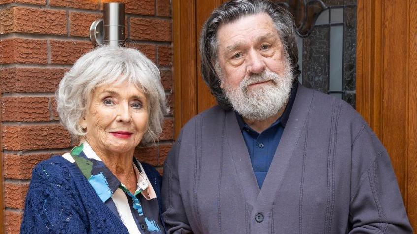 Sue Johnston and Ricky Tomlinson are pictured in character, standing in front of a semi-detached red-bricked house on Brookside Close. A wooden door with a glass panel is behind them and a window is to the left. Johnston is wearing a colourful patterned dress and a navy cardigan and Tomlinson is wearing a navy polo shirt and dark grey cardigan.