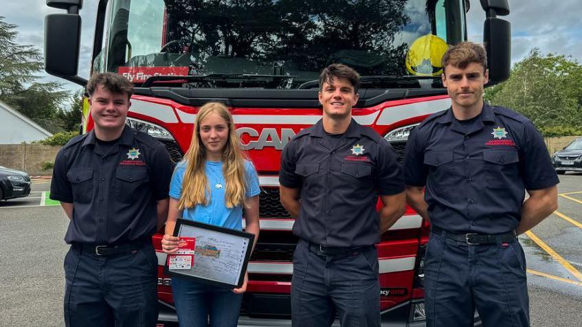 Three firefighters in blue short-sleeved shirts and trousers, standing with Harriet, who has long, fair hair and is wearing a blue T-shirt and holding her hand-drawn design. All four are standing in front of a bright red and white striped fire engine. 