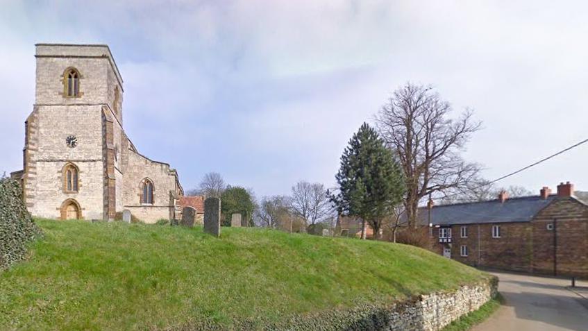 Pattishall church on a grassy mound alongside a village road and houses