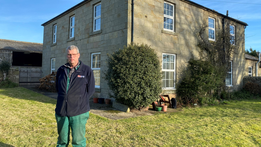 Martin Beal, a man in his 50s, is standing outside his house. He is wearing a blue top and green trousers.