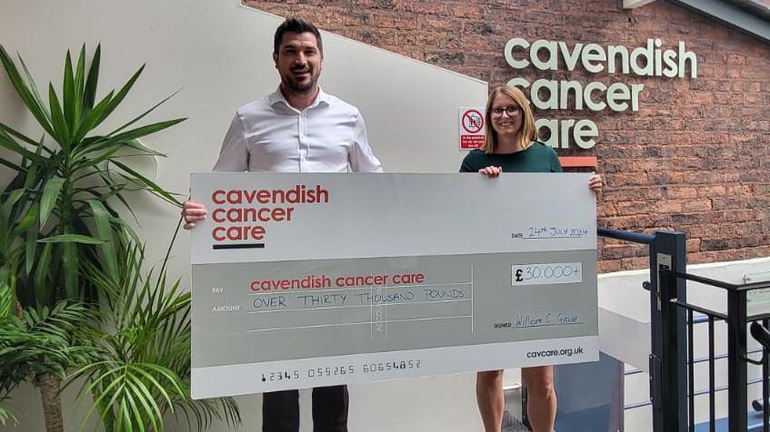 Liam and a woman are holding a giant cheque in Cavendish Cancer Care's building. It reads: "Pay Cavendish Cancer Care thirty thousand pounds, signed William C Graves."