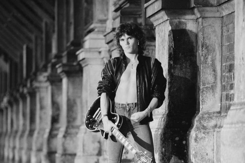 A black and white image of Robin George. He is wearing jeans and a black leather jacket, carrying a guitar. 