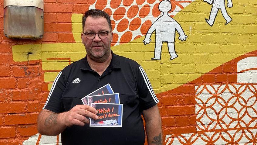 A man wearing glasses and a black Adidas t shirt holds postcards with  Wish I wasn't here written on them. The cards also depict someone sleeping in a doorway. He is standing in front of a brightly painted orange and yellow wall