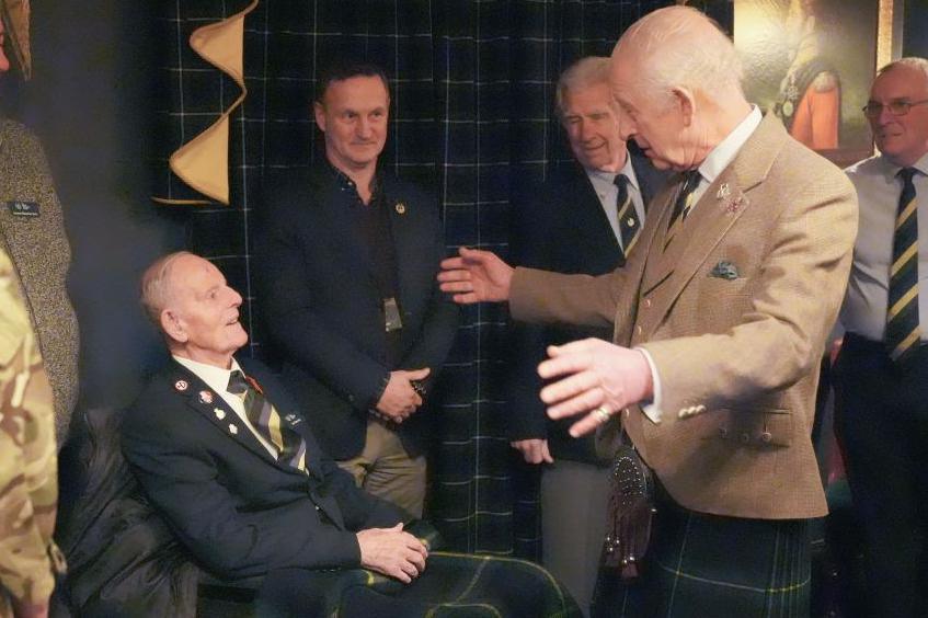 Jim Glennie, 99-year-old man wearing a striped tie and a jacket, speaking to King Charles in a light brown jacket and kilt, with his arms outstretched