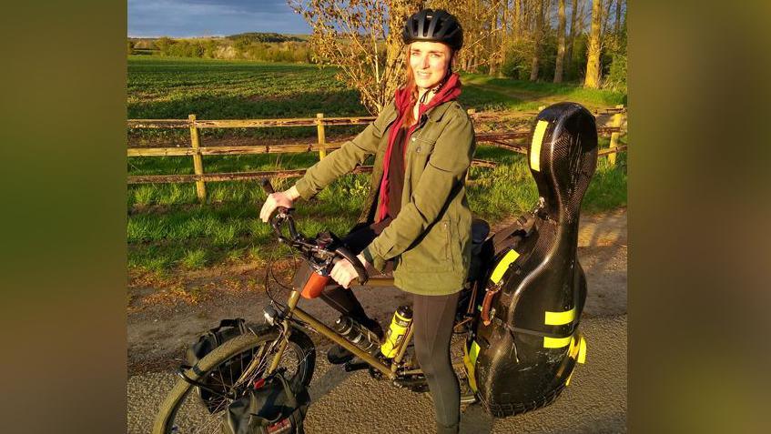 A woman in a green coat on a black bike with a large black instrument case. She is posing by a field.