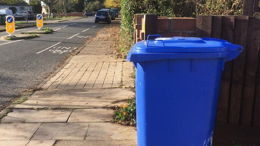 A blue recycling bin sits on a pavement outside a property. A road can be seen nearby with cars parked on it.