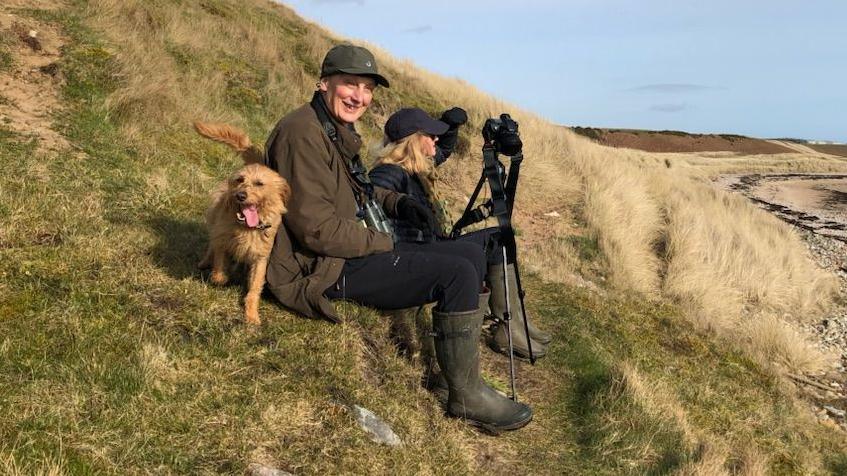 Prof Crellin sat on a hillside with his wife and dog with a camera on a tripod in front of him 