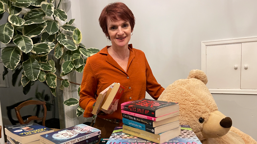 Helen is standing behind a table filled with books and games as well as a huge teddy bear. She is wearing a russet-coloured blouse and is smiling to camera, holding several books in her hand