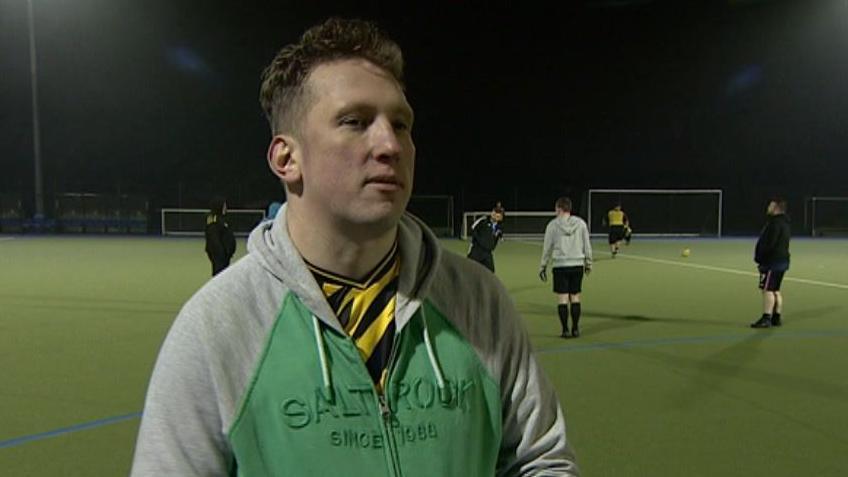 Joe Evans, a man with short brown hair, is wearing a green and grey hoodie with a yellow and black striped football shirt underneath. He is stood on a football pitch, with a group of players behind him.