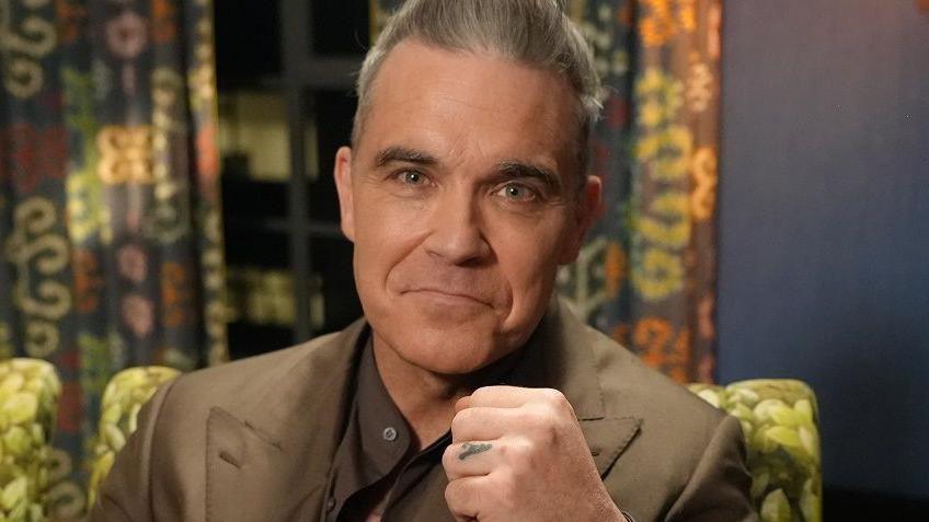 Robbie Williams pictured smiling whilst raising a fist to the camera. He has short greying hair, pulled back from his forehead, and his wearing a light brown suit over a dark brown collarless shirt. 