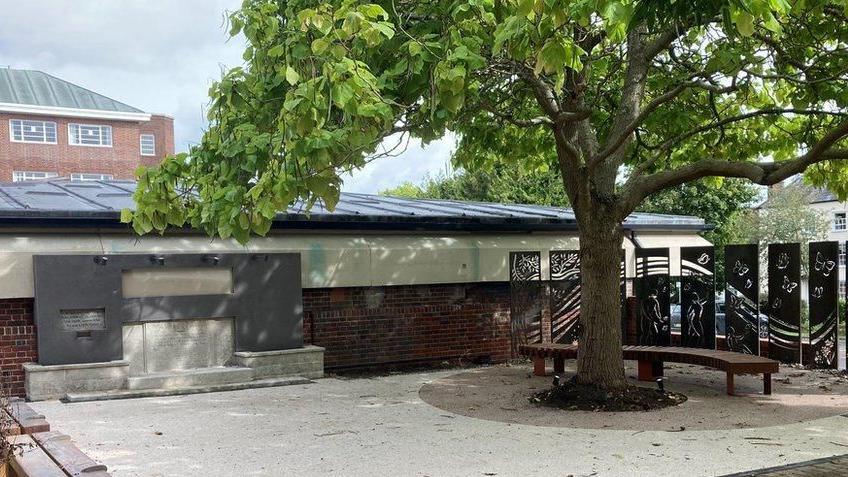 A white paved area in front of a brick wall with a mature tree and bench on the right and a stone memorial on the left. The memorial is flat against the brick wall and looks roughly