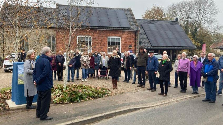 A small group of people stand together looking toward Kenneth and another woman. The Pulham Airfield marker can be seen under a blue sheet prior to it being unveiled.