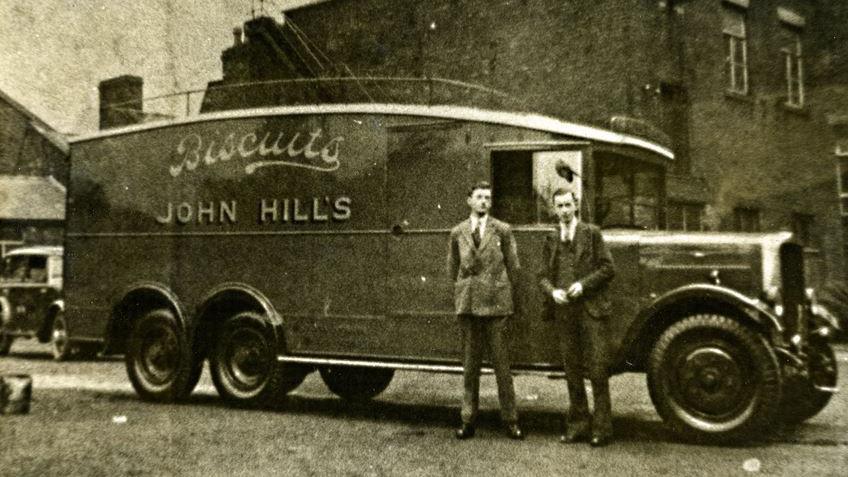Two men in period suits stand in front of a Hill's Biscuits delivery van in a black and white picture taken in the 1940s.