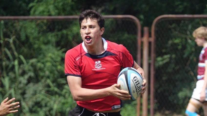 Seb Pena wearing a red jersey has a rugby ball in his hand. He appears to be in motion, and there is a rusty fence behind him.