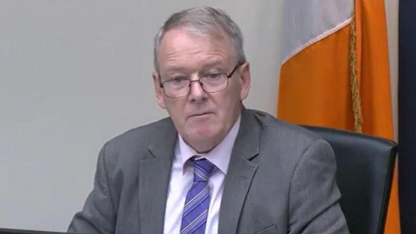 Brian Stanley is sitting in a committee. He's wearing black glasses and a grey suit with a white shirt and a purple tie. The Irish tricolour flag is behind him.