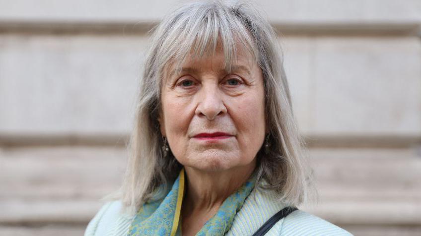 A woman with shoulder length greying hair and a pale green jacket stares directly at the camera