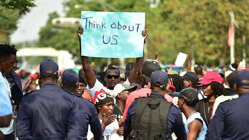 A protester holds a placard that says "think about us" in English as demonstrators gather in Luanda. Some policemen with their backs to the camera can be seen in the foregound.