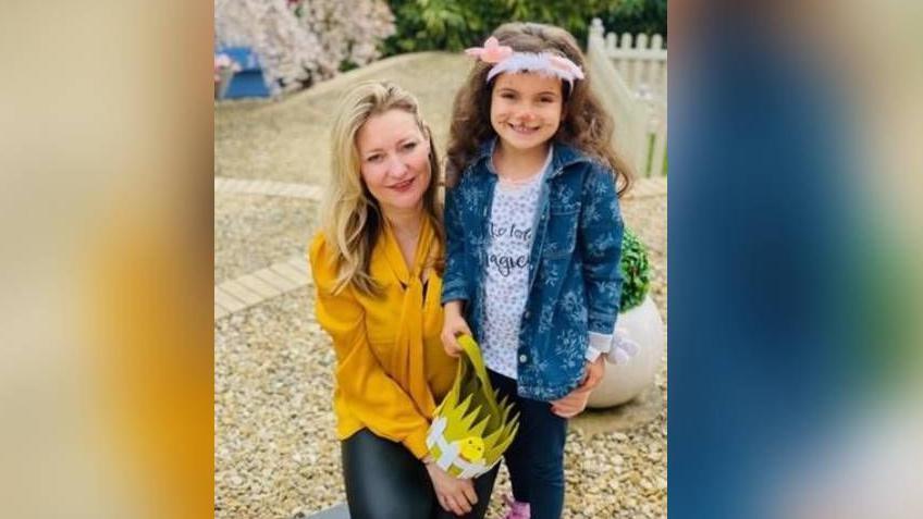 A woman who has blonde hair wearing a yellow top. She is holding a young girl around her waist as the pair smile for a photo. The girl has cat face paint on her face and is wearing a blue denim jacket and white top. They are stood in a stone area.