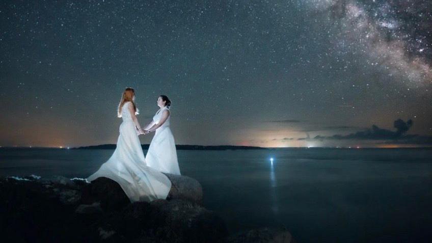 Two brides hold eachother's hands while standing on a rock near the sea across which is a landmass. It is night-time and the Milky Way is illuminated and stars are shining.