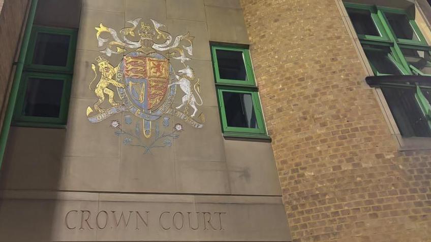 Luton Crown Court: The words "CROWN COURT" are written below a multi-coloured crest on a grey wall above an entrance to a brown-brick building with green-framed windows.